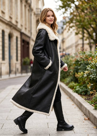Side view of a model walking in a women's black leather and faux shearling longline trench coat paired with black boots.