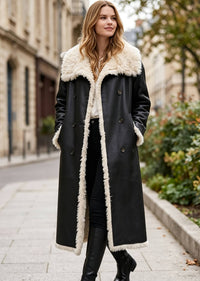 Model wearing a double-breasted black faux leather coat with plush shearling lapel collar and trim, hands in pockets.