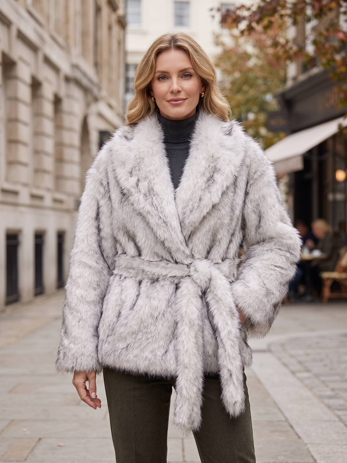 A woman wearing a light gray faux fur wrap coat with a tie belt, styled over a black turtleneck and dark olive pants, standing on a city street.