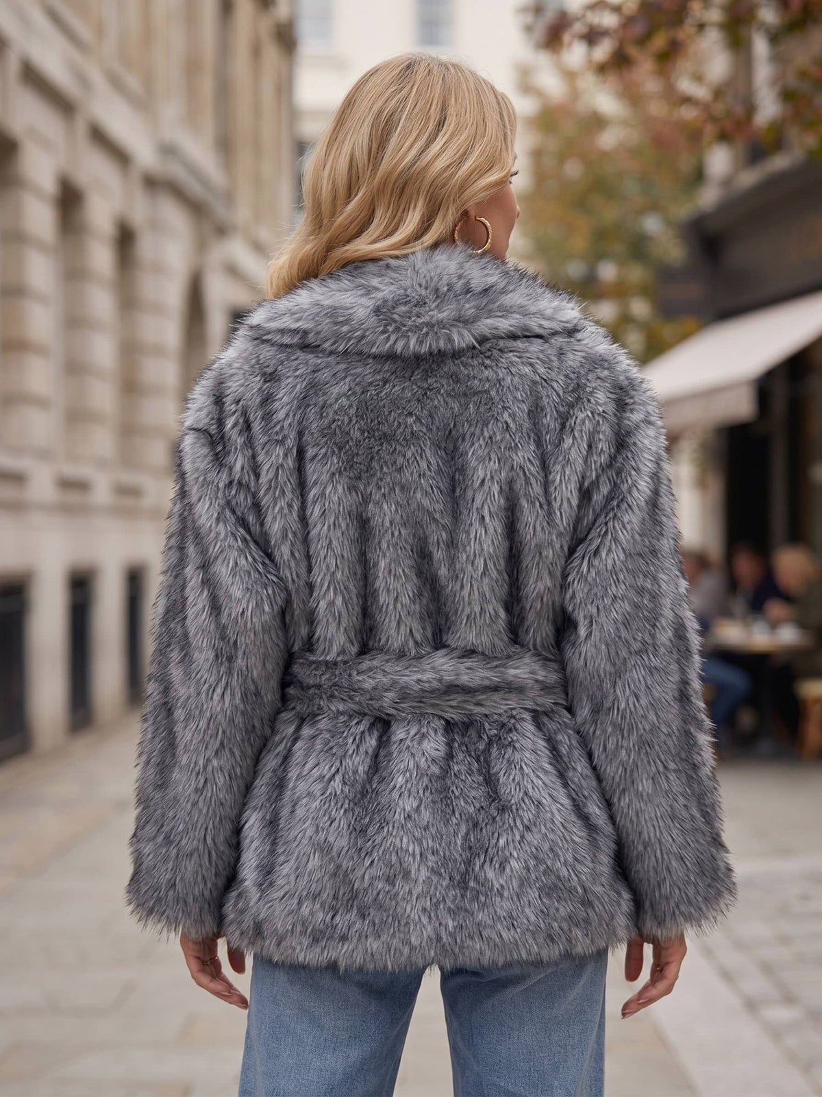 A woman viewed from behind wearing a gray faux fur belted coat and blue jeans, standing on a city street.