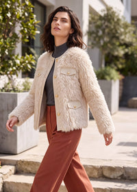A woman walking outdoors wearing a cream faux-fur cropped jacket with gold-tone buttons and flap pockets over a dark gray top and rust-brown wide-leg pants.