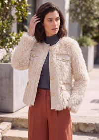 A woman wearing a cream-colored, shaggy faux-fur cropped jacket with gold-tone buttons and flap pockets, styled over a dark gray turtleneck and rust-brown wide-leg trousers, standing outdoors.