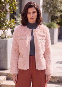 A woman wearing a light pink shaggy faux-fur jacket with gold buttons, styled over a dark gray turtleneck and rust-brown trousers, standing outdoors in front of a modern building.