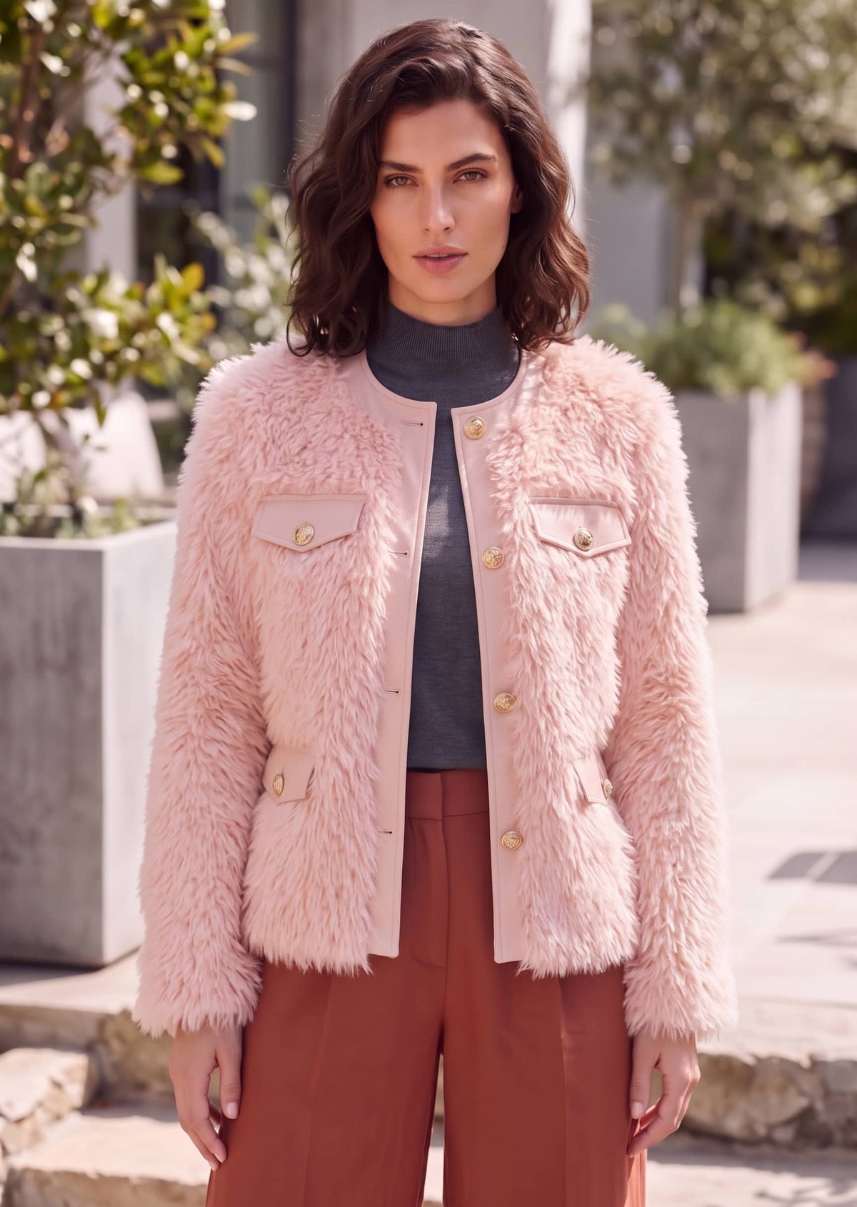 A woman wearing a light pink shaggy faux-fur jacket with gold buttons, styled over a dark gray turtleneck and rust-brown trousers, standing outdoors in front of a modern building.
