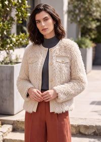A woman wearing a cream faux fur jacket with gold buttons over a dark gray turtleneck and rust-colored pants, standing outdoors in front of a modern building with greenery.