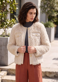 A woman wearing a cream faux fur jacket with gold buttons, paired with a black turtleneck and rust-colored trousers, standing outdoors in front of a modern building.