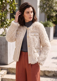A woman wearing a cream faux fur jacket with gold buttons over a dark gray turtleneck and rust-colored wide-leg pants, standing outdoors in front of modern buildings and greenery.