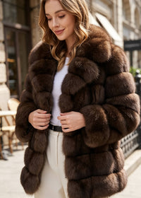 A woman wearing a luxurious dark brown fur jacket with a wide collar, styled over a white top and cream trousers, standing on a city street.