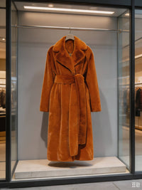 Display window view of a plush rust orange belted faux rabbit fur long coat hanging inside a modern boutique storefront.