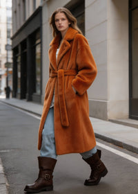  Side profile shot of a blonde model striding forward in a long rust orange faux fur wrap coat with a tied waist belt.
