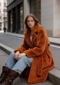 Seated portrait of a woman wearing a plush rust velvety faux rabbit fur long coat with wide lapels on an urban curb.