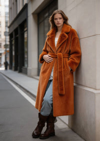 Model leaning against a building wall wearing a long velvety rust-colored faux rabbit fur coat with a matching waist tie.