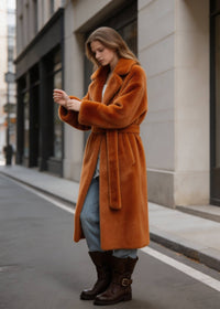 Side view of a woman looking down and adjusting the sleeve cuffs of a rust-toned velvety long faux fur coat.