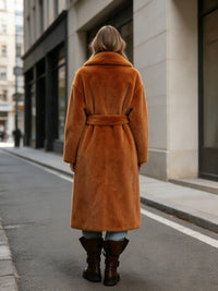 Full back view showing the rear fit and length of a women's rust brown belted long faux rabbit fur coat outdoors.