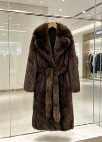 A long brown fur coat with a wide collar and matching belt, hanging on a wooden hanger inside a glass display case in a boutique.