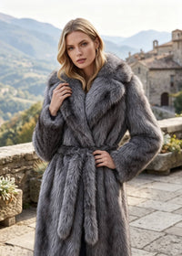 A woman wearing a luxurious gray fur wrap coat with a matching belt, standing on a stone terrace with a scenic mountain and village background.