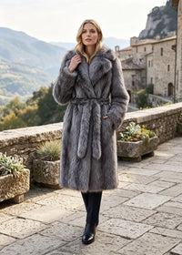 Full-length shot of a woman modeling an ash grey faux fur belted trench coat with dark trousers and black boots on a stone patio.
