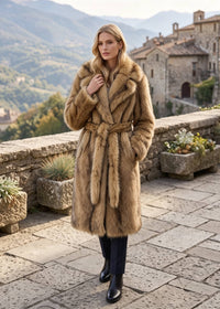Front view of a blonde woman walking outdoors in a camel brown belted faux fur wrap trench coat.
