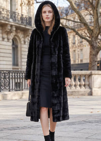 A woman wearing a long black faux fur coat with a hood over a black dress, standing outdoors on a stone walkway with a city building in the background.