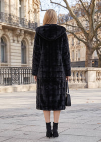 Back view of a woman wearing a black longline tiered faux fur coat with an oversized hood and black heeled ankle boots on a city street.