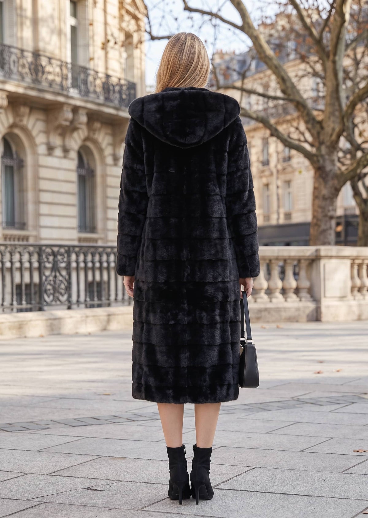Back view of a woman wearing a black longline tiered faux fur coat with an oversized hood and black heeled ankle boots on a city street.