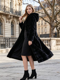 Side movement shot of a woman modeling a flowing black longline hooded faux fur coat over a tailored black knee-length dress.