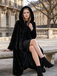 Elegant seated pose of a blonde woman wearing a black paneled faux fur winter coat with the hood pulled up on an outdoor stone bench.