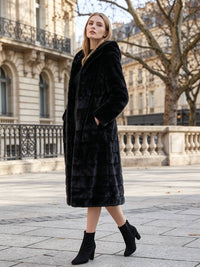 Three-quarter profile shot of a model standing with hands in pockets wearing a warm black hooded faux fur coat in an urban Parisian setting.