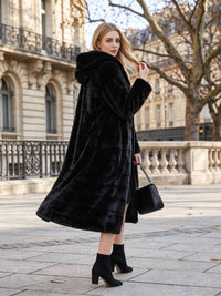 Street style fashion shot of a woman looking over her shoulder wearing a flared black tiered faux fur coat with a hood and holding a leather handbag.
