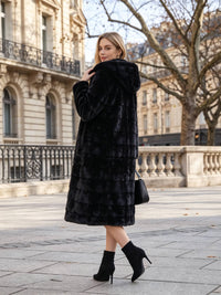 A woman wearing a long black faux fur coat with a hood, black ankle boots, and carrying a black handbag, posing outdoors on a city street with classical stone buildings in the background.