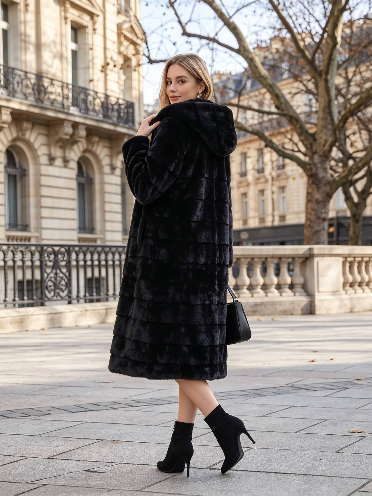 A woman wearing a long black faux fur coat with a hood, black ankle boots, and carrying a black handbag, posing outdoors on a city street with classical stone buildings in the background.