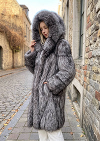Three-quarter view of a woman wearing a silver-gray fox faux-fur hooded longline winter coat with the hood pulled up, looking downward in an outdoor alley.
