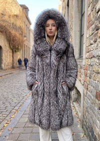 Front view of a woman wearing a longline silver-gray fox faux-fur winter coat fully zipped with the large hood up, hands in pockets on a cobblestone street.