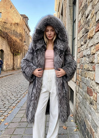 Front view of a blonde woman wearing an open silver-gray fox faux-fur longline winter coat with an oversized hood, paired with a pink top and white wide-leg sweatpants on a cobblestone street.