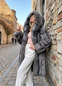A woman wearing a luxurious long gray fur coat with a large hood, styled over a light pink top and white pants, posing on a cobblestone street next to a rustic stone wall.