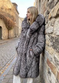 Side profile of a woman leaning against an exterior brick wall, wearing a thick silver-gray faux-fur longline winter coat with a plush oversized hood.