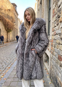 Front three-quarter view of a blonde woman wearing a hooded silver-gray fox faux-fur coat draped open at the front over casual clothes on a cobblestone path.