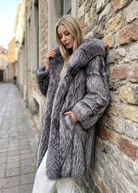 Side view of a blonde woman leaning against a rustic stone wall, holding the lapel of a heavy silver-gray fox faux-fur hooded longline coat.