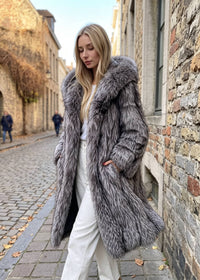 hree-quarter view of a woman walking down a cobblestone street, wearing an open silver-gray fox faux-fur longline coat over a white top and wide-leg trousers.