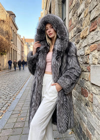A woman wearing a long, luxurious gray and black fur coat with a large hood, styled over a light pink crop top and white drawstring trousers, standing on a cobblestone street next to a stone wall.