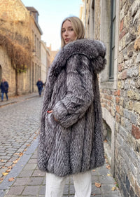 Over-the-shoulder side view of a woman wearing a voluminous silver-gray fox faux-fur longline winter coat with an oversized hood.