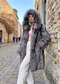Full-length view of a woman leaning against a stone wall, modeling an open silver-gray fox faux-fur longline coat paired with a crop top and white trousers.