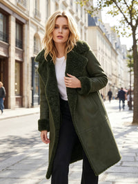 A woman stands on a city street wearing a long olive green coat with a plush fuzzy collar and cuffs, layered over a white top and dark pants.
