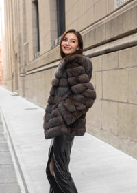 A three-quarter view of a woman smiling and looking back, showcasing the texture and volume of her hooded brown fur coat and black leather skirt.