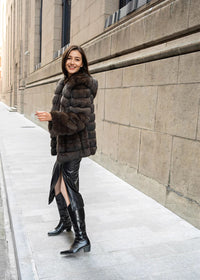 A full-length shot of a woman looking back over her shoulder with a smile, wearing a thick brown fur coat, a slit black leather skirt, and tall black leather boots.