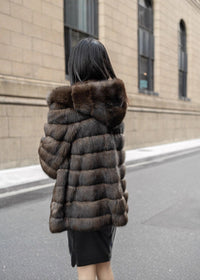 A view from behind of a woman with long dark hair walking down a city street, wearing a hooded, hip-length brown fur coat with horizontal paneling.