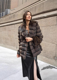A woman looking off-camera while posing in a textured brown fur coat, a grey top, and a long black leather skirt with a high leg slit.
