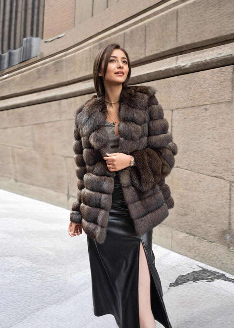 A woman looking off-camera while posing in a textured brown fur coat, a grey top, and a long black leather skirt with a high leg slit.
