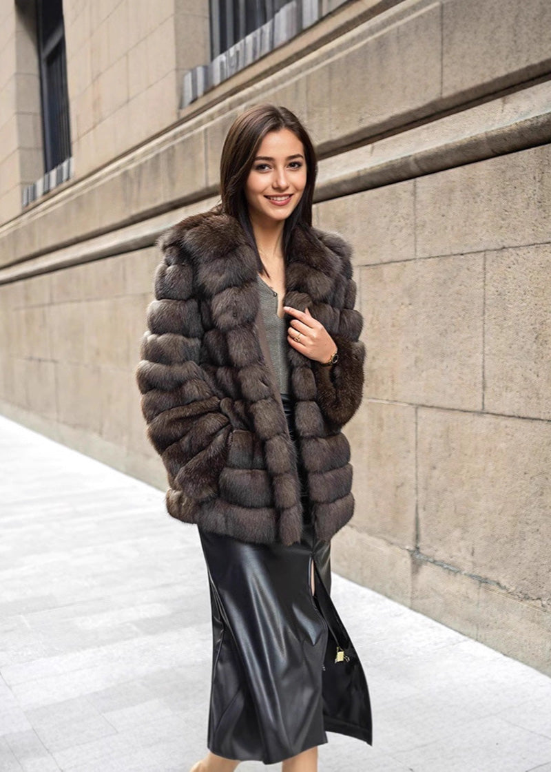 A woman posing while walking forward on a stone sidewalk, wearing a luxurious brown fur coat over a grey shirt and a black leather skirt with a slit.