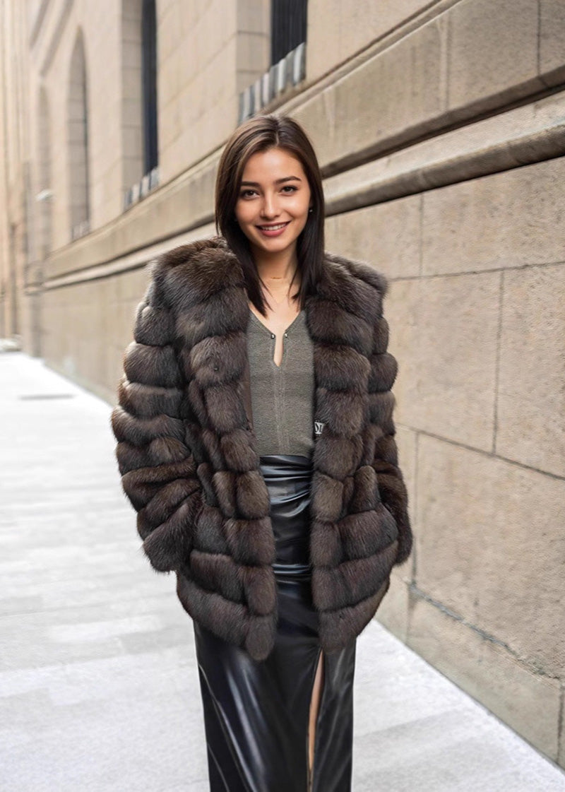 A medium shot of a smiling woman standing on a city sidewalk, wearing an open brown fur jacket over a grey top and a black leather skirt.